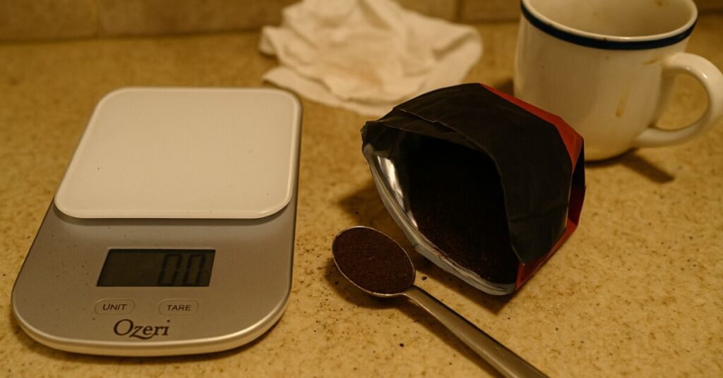 Digital coffee scale with ground coffee, measuring spoon, and open coffee bag on a cluttered kitchen counter with stained mug and paper towel, illustrating home drip coffee brewing setup